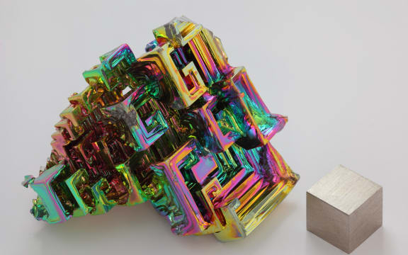 The chemical element bismuth as a synthetic crystal. The iridescent surface is a very thin layer of oxidation. Beside it is a high purity (99.99 %) 1 cm3 cube of bismuth for comparison.