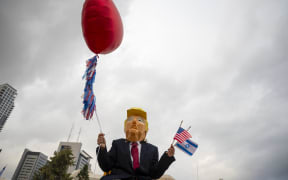 A person wearing a mask depicting US President Donald Trump holds US and Israeli flags after the hostage deal declared by Trump, at Hostage Square in Tel Aviv.