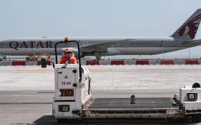 (FILES) In this file photo taken on April 1, 2020, an airport worker wearing a face mask (protective measure during the COVID-19 coronavirus pandemic) mans a luggage trolley while behind him is seen a Qatar Airways Boeing 777 aircraft at Hamad International Airport in the Qatari capital Doha.