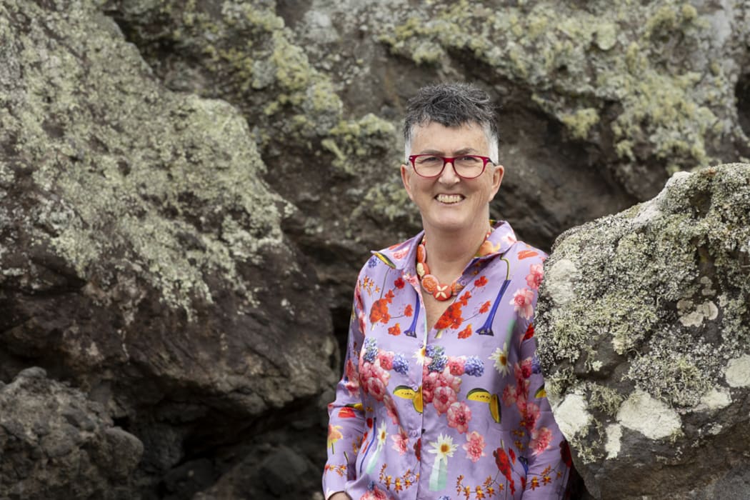 Dr Lucy O'Hagan - a woman with short grey hair and red glasses - stands smiling between some large rocks.