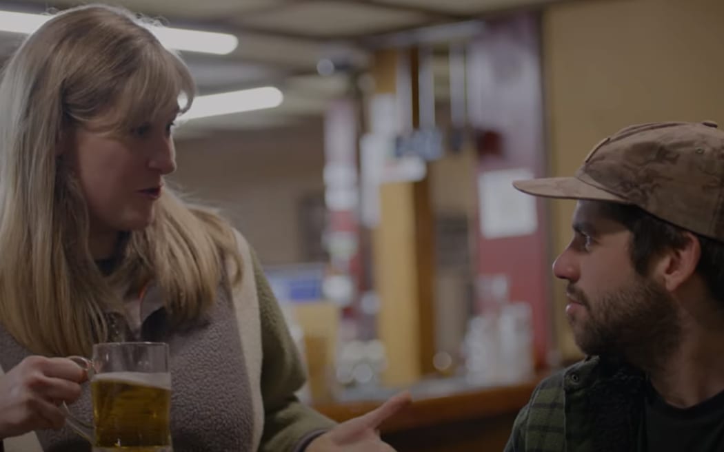 Brynley Stent, a white woman with blonde hair, holds a beer and speaks with a seated dark-haired man in cap.
