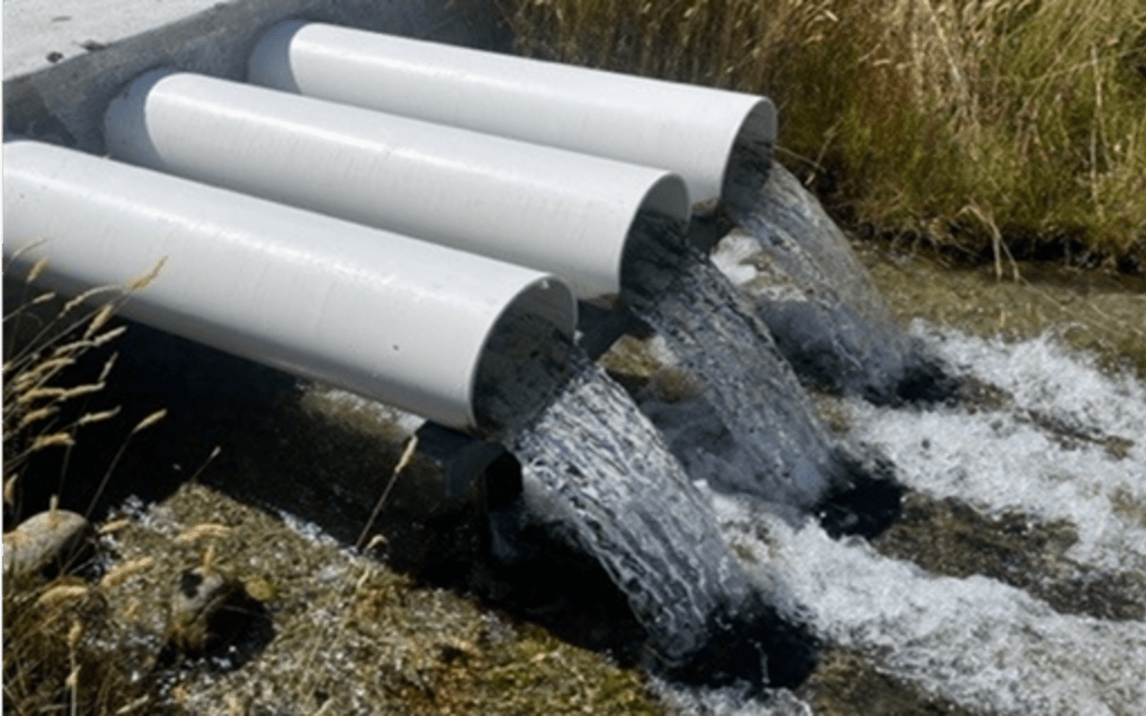 A concrete barrier across a stream with three pipes gushing with water. The surrounding vegetation is grass and tussock.