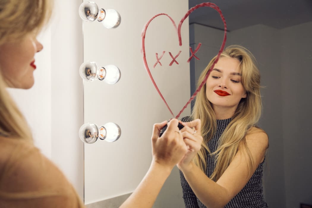 A woman looking in a bathroom mirror drawing a heart on the mirror with lipstick. (Photo by Manuela / Cultura Creative / Cultura Creative via AFP)