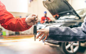 Car workshop, woman picking up her car - Mechanic working on car engine, woman giving automobile key for a check up