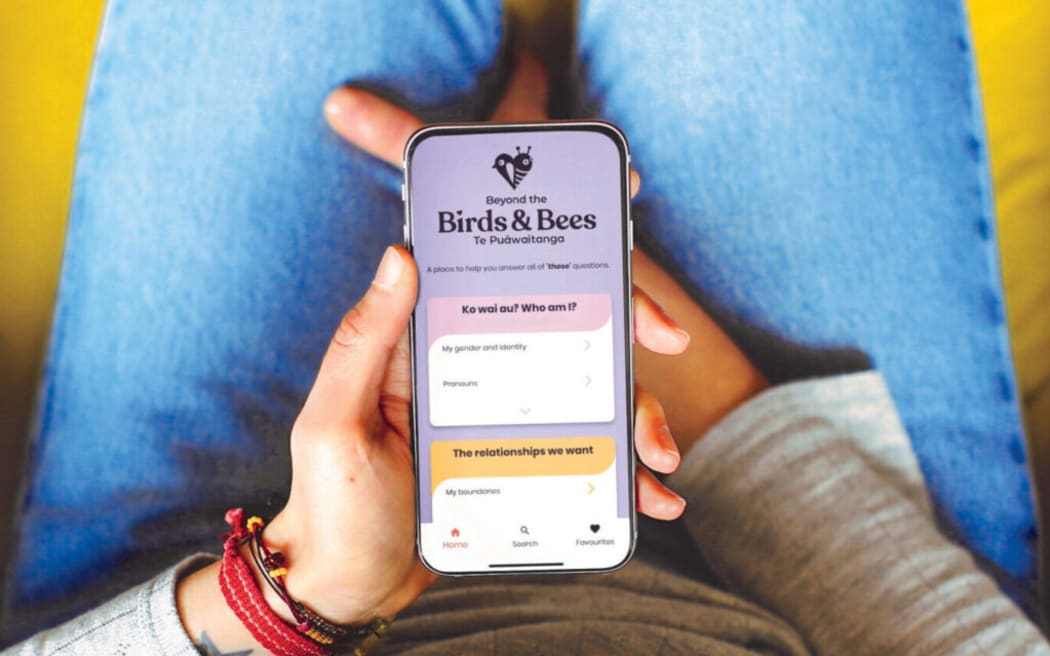 A young person holds a phone with the screen showing the Te Puāwaitanga: Beyond the Birds & Bees app
