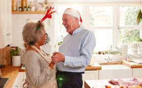 Loving Senior Couple Wearing Fancy Dress Antlers Dance In Kitchen Whilst Preparing Christmas Dinner