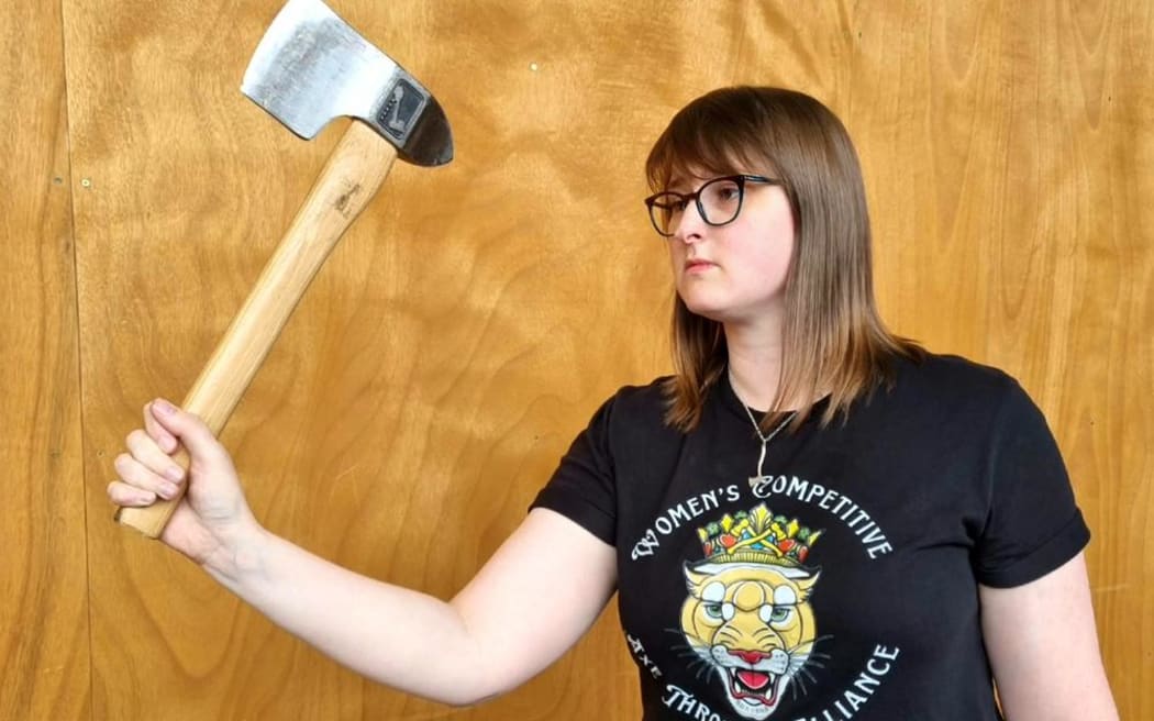 Britt holds an axe and looks poised to throw it. She is standing in front of a wooden wall and wearing a black tshirt.