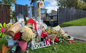 A pile of flower bouquets lay on the side of the road, on grass, and a letters spell out aroha.