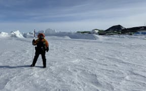 A person wearing black and orange cold-weather gear with the hood up stands on a flat expanse of ice criss-crossed with weaving patterns. In the background, jumbles of ice pile up. The buildings of Scott Base and black hills are visible in the background on the right, beneath a cloud-streaked blue sky.