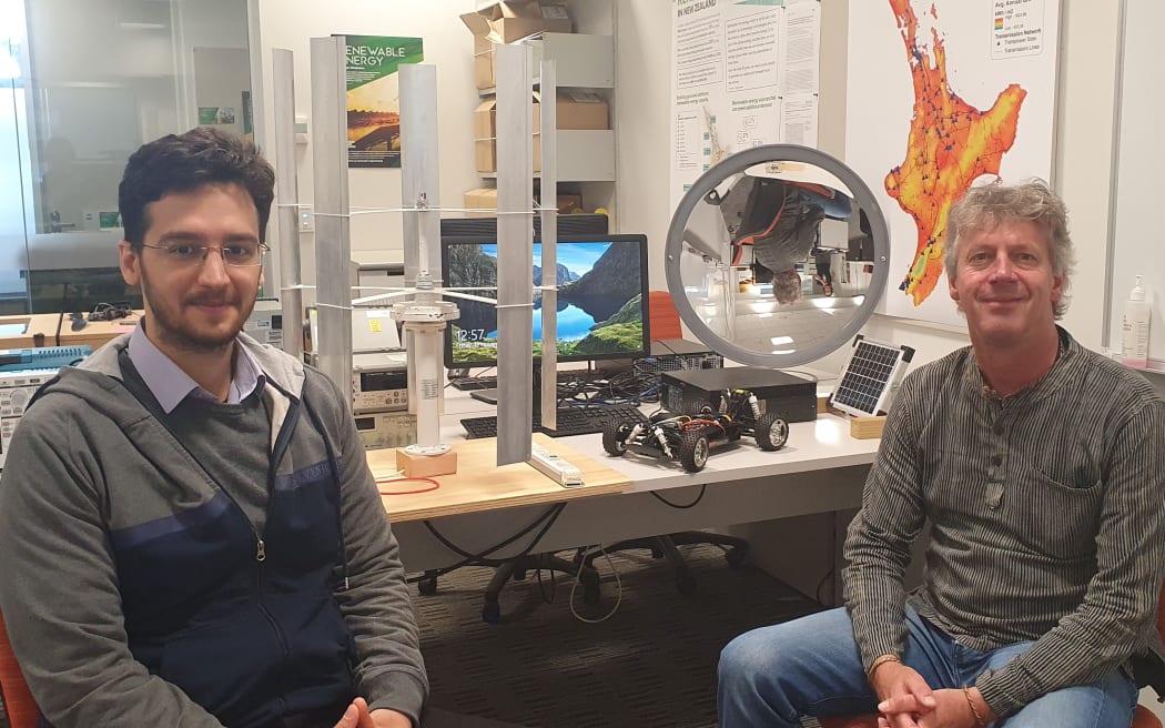 Soheil Mohseni and Alan Brent work with sustainable energy systems. There are mini versions of a vertical wind turbine and a solar concentrator on the desk behind them.