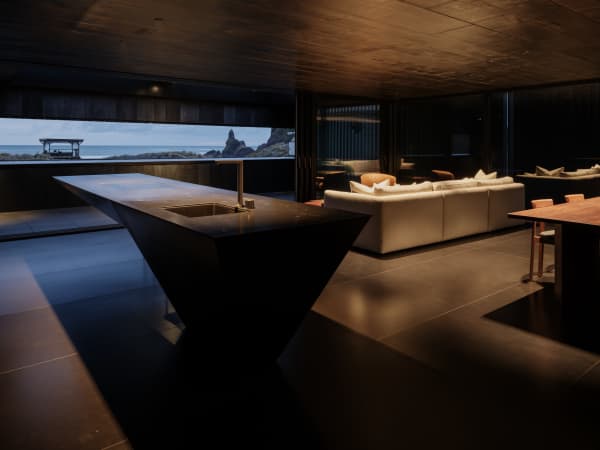 The dark living room interior of Bunker House in Piha, with the window framing a view of the coastline.