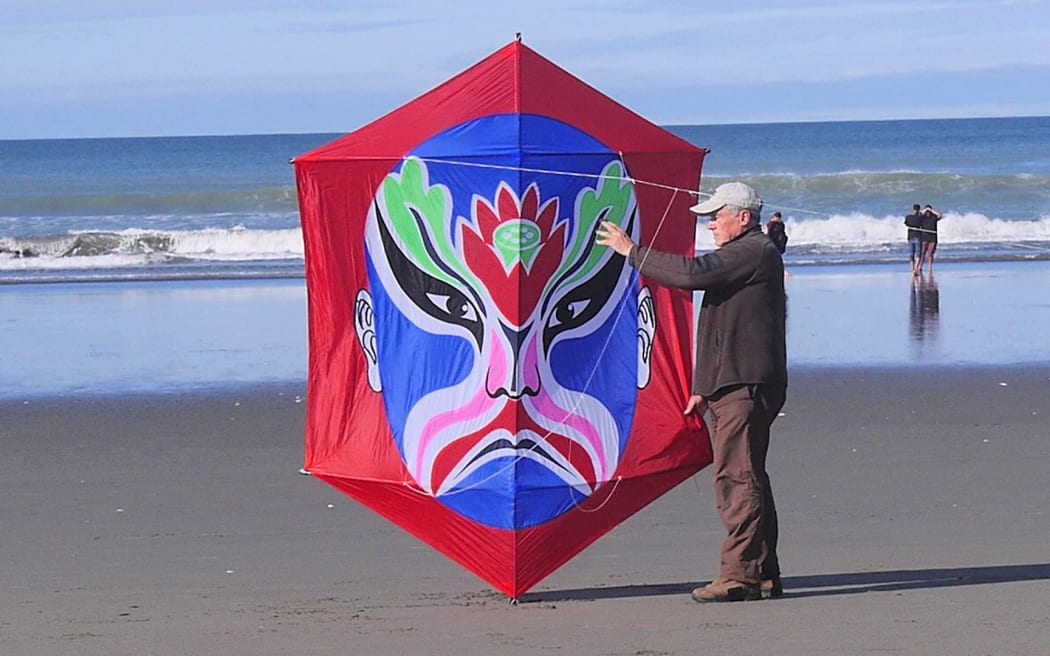 Jim Nicholls stands on the beach holding a giant red kite with a rokkaku design on it.