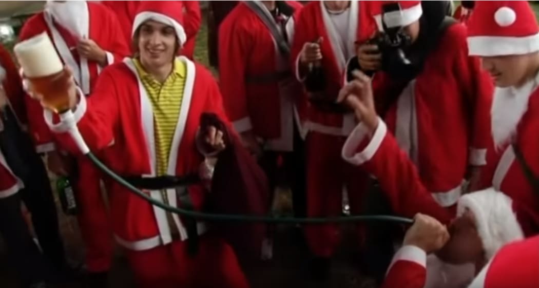 A Santa drinks from a beer funnel held by another Santa.