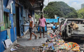 This frame grab taken from AFPTV video footage taken on January 11, 2024 shows people walking past debris in front of buildings damaged during riots in Port Moresby. Papua New Guinea's prime minister declared a 14-day state of emergency in the capital on January 11, after 15 people were killed in riots as crowds looted and burned shops. (Photo by Darrell Toll / AFPTV / AFP)
