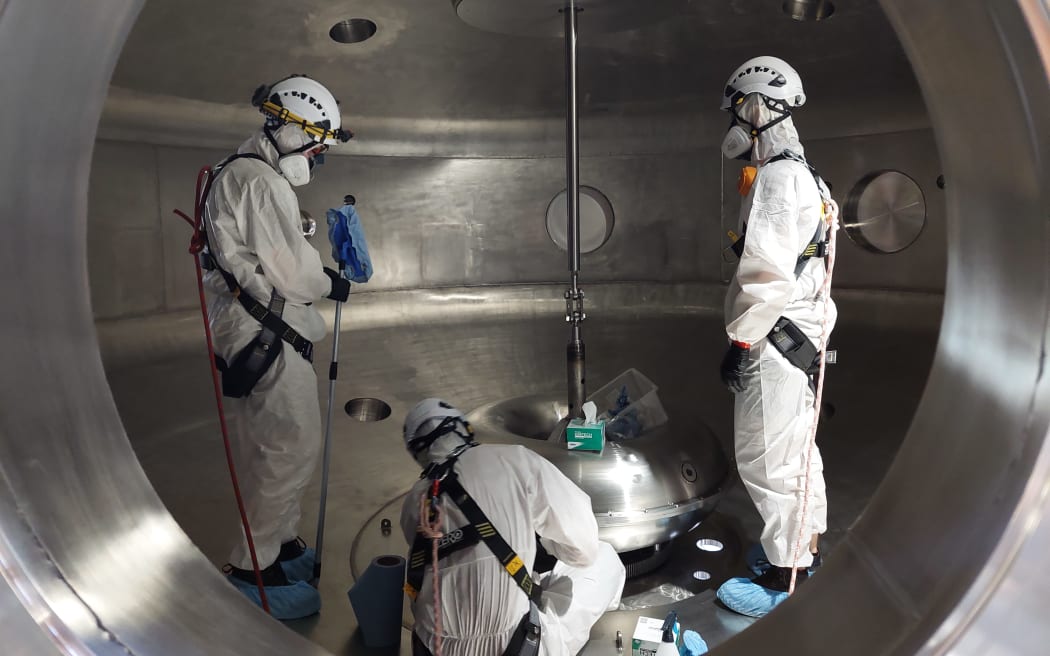 Three people wearing white jumpsuits, harnesses, helmets and respirators inside a metal chamber, gathered around a large metal donut object that has a metal pole extending from the chamber's ceiling through its centre. They are viewed through a circular opening.