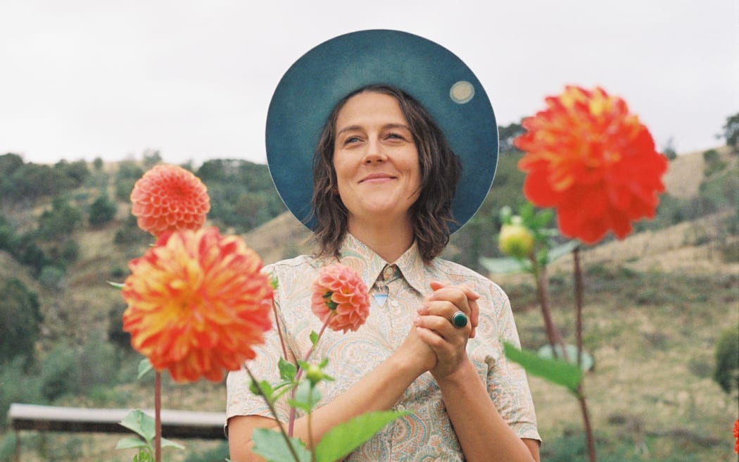 A photo of country musician Kerryn Fields standing in a field with red flowers in front of her, Kerryn is wearing a round brim hat and smiling.