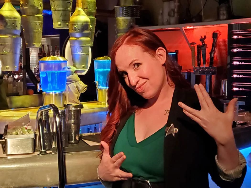 Erin stands in front of a sci-fi set. She is wearing a Star Trek uniform. She smiles and performs the Klingon peace sign for "live long and prosper".