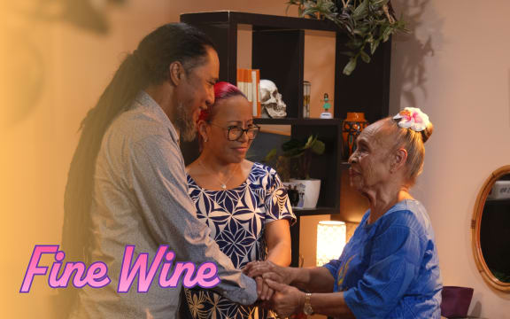 A man with dreadlocks holds hands with an older woman wearing a flower in her hair. Text reads "Fine Wine".