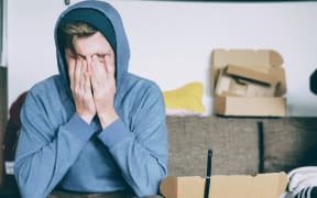 A man wearing a light blue hoodie sits on a brown sofa with his hands obscuring his face, surrounded by boxes.