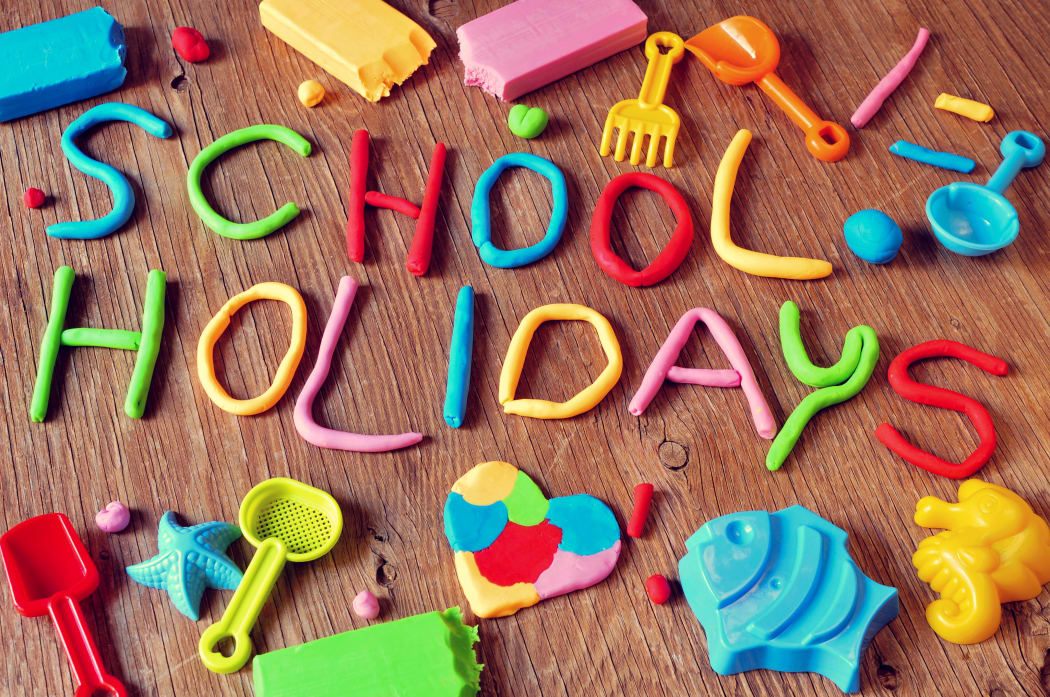 the text school holidays made from modelling clay of different colors and some beach toys such as toy shovels and sand moulds, on a rustic wooden surface