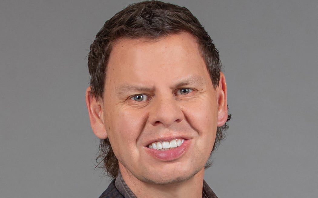 A brown-haired man in a checked jacket looks at the camera, smiling slightly