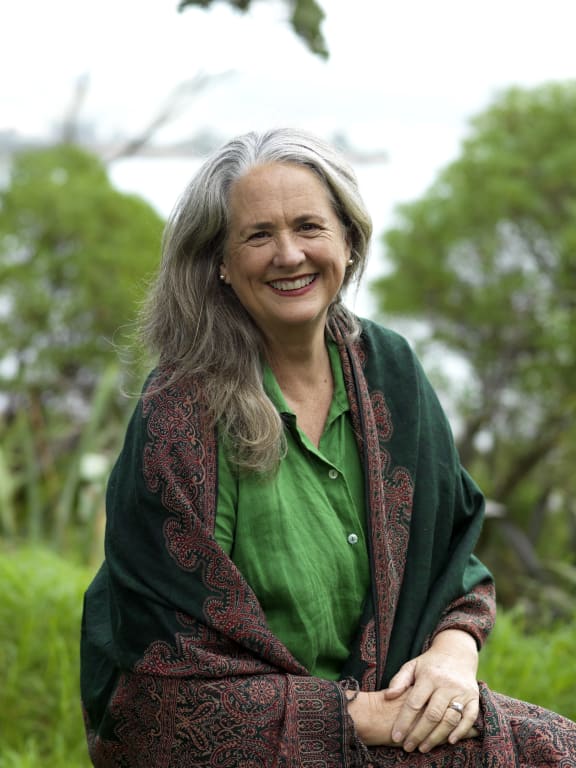 Wendyl smiles at the camera. She is outside in a clearing surrounded by lush bush. The sea is in the distance. It is a bright, pale day.
