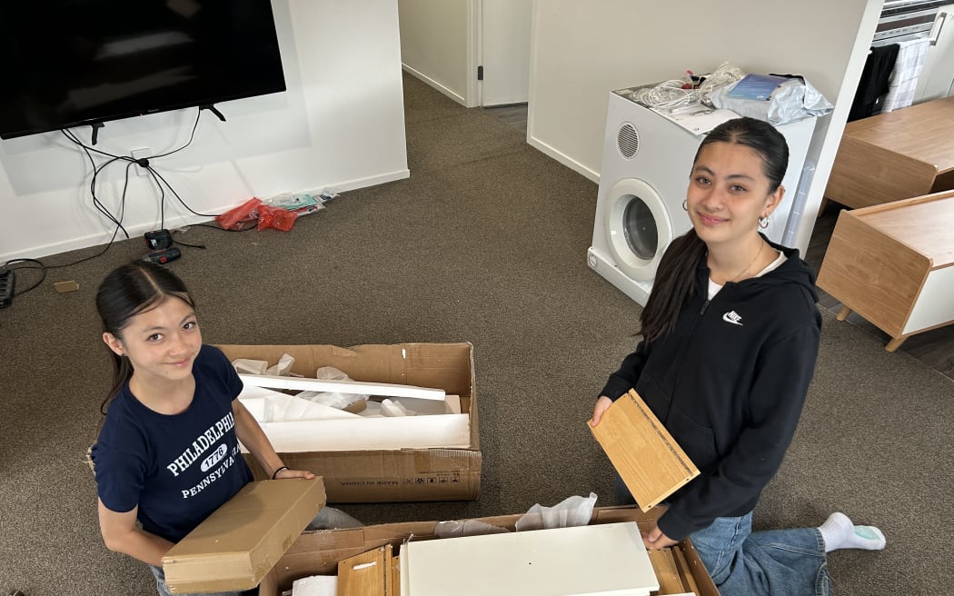 Picture of two young women smiling at camera, sitting among furniture pieces.