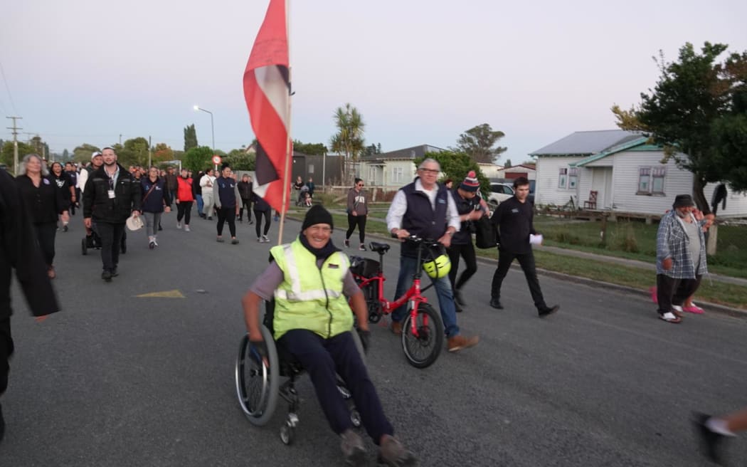Wairoa resident Val Irwin is in a wheelchair and wears a yellow hi vis vest and black beanie. He has a tino rangatiratanga flag attached to the back of the wheelchair and other people walk alongside and behind him.