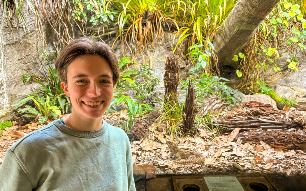 A person wearing a grey-green crewneck sweater stands in front of the glass of a zoo exhibit. The enclosure is filled with leaf litter, ferns, and rocks, and a grey-green reptile that looks like a lizard (but isn't) sits in the leaf litter.