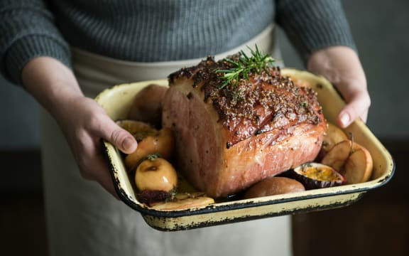 A person wearing an apron holds a casserole of baked ham with roasted apples on the side.