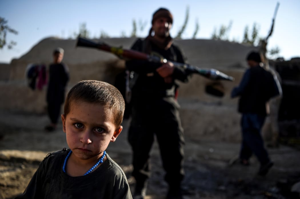 A young boy looks at the camera as a policeman holding a rocket-propelled grenade stands behind in a house at Deh Qubad village in Maiwand district of Kandahar province on September 27, 2020.