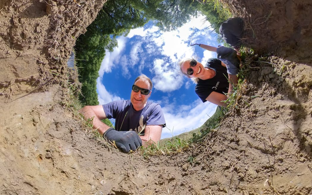Kirstin and Matt peer into a hole in the ground, smiling.