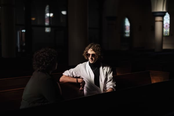Presenter Ari Kerssens is seated in a church wearing a white shirt and glasses. He is surrounded by darkness.