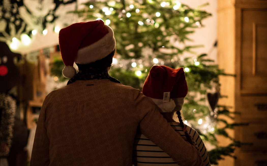 An adult and a child wearing Christmas hats sitting in front of a Christmas tree with their backs to the camera.