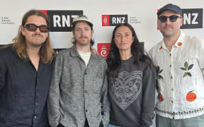 Three members of the band Leisure stand to have their picture taken with Kara Rickard in front of the RNZ wall.