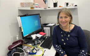 A woman sits at her desk in a medical office. The desk is strewn with papers, equipment, with a computer desktop sitting in the centre. The woman is facing the camera in her chair, smiling.