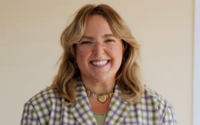 Lucu Blakiston, a women in her later 20's with shoulder length blonde hair, smiles at the camera. She wears a checkered button up top.