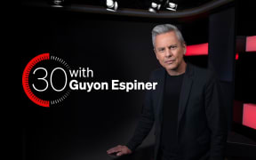 Guyon Espiner on a black background, looking at camera with logo behind him