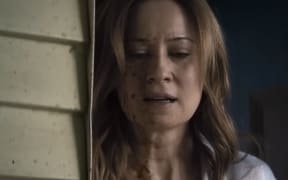 A long-haired woman with a blood-spattered face stands in a doorway looking shocked.