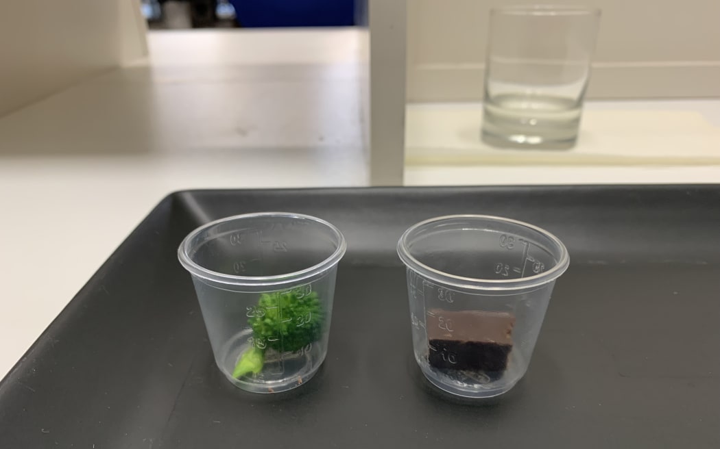 On a black tray two small clear plastic cups sit side by side. One contains a sprig of broccoli and the other a square of dark chocolate.