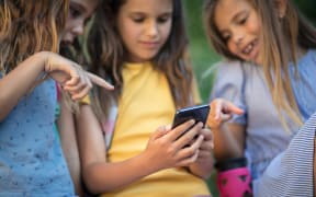 Three young girls talking and pointing at one of their phone screens.