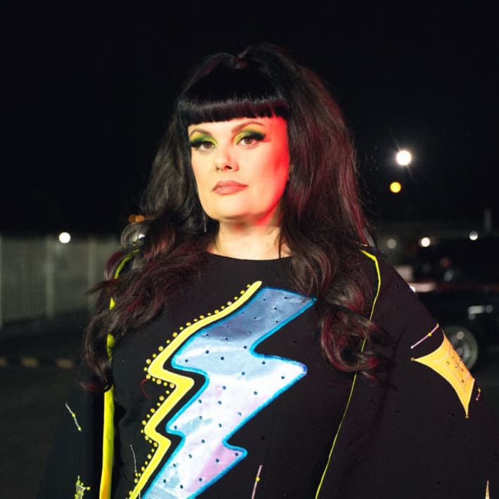 A woman with long black hair wearing a black top with a colourful lightning bolt gazes at the camera.