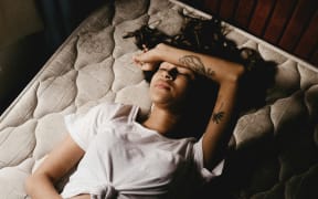 A woman reclines on an unmade bed, with one forearm across her forehead, the other on her stomach.