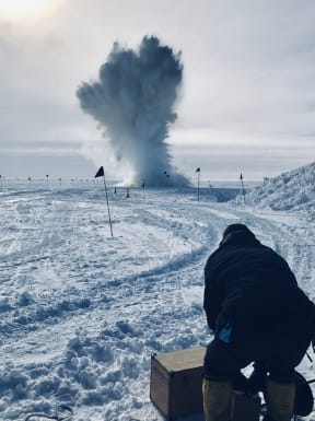 A scene on an icy expanse with vehicle tracks indicated by flags on poles. An explosion in the distance seeds a cloud of snow and ice jetting into the air. A person crouches in the foreground with their back to the camera, watching the explosion.