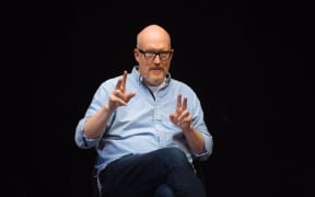 Joe stands on a stage gesturing and speaking against a black background.