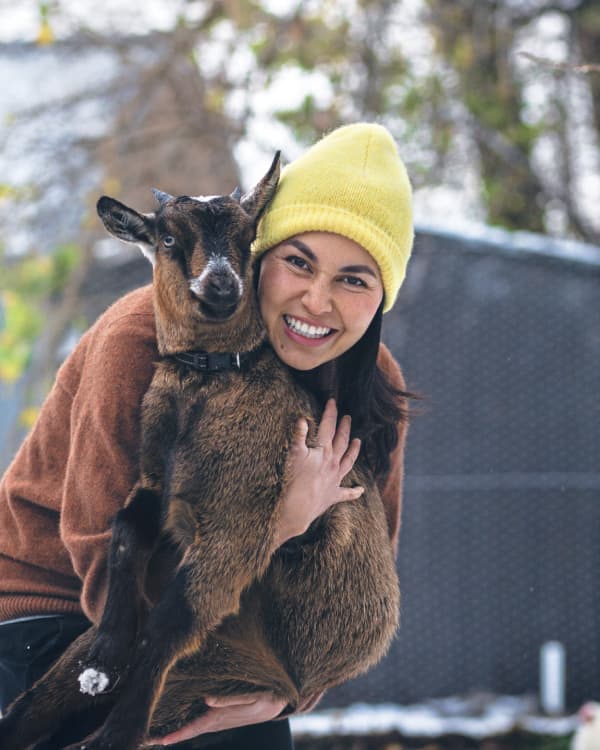 Nadia Lim smiles and hugs a goat while wearing a bright yellow beanie.