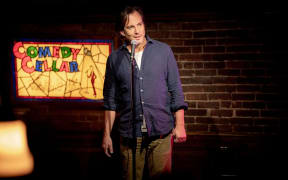 Will Arnett, wearing a partially buttoned blue shirt, talks into a standing microphone with the words 'Comedy Cellar' on an illuminated sign behind him.