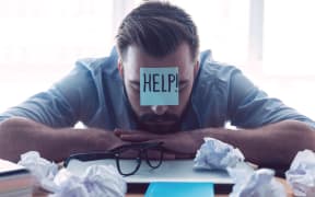41257198 - he needs help. hopeless young beard man with adhesive note on his forehead leaning at the desk and keeping eyes closed while sitting at his working place in office