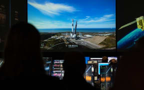 A group of people in a control room watch a large screen displaying a rocket on a launch pad, with a countdown timer at T-00:00:01. The screen also shows telemetry data and an image of Earth with a satellite. Multiple computer monitors in the control room display various data and graphs related to the rocket launch.