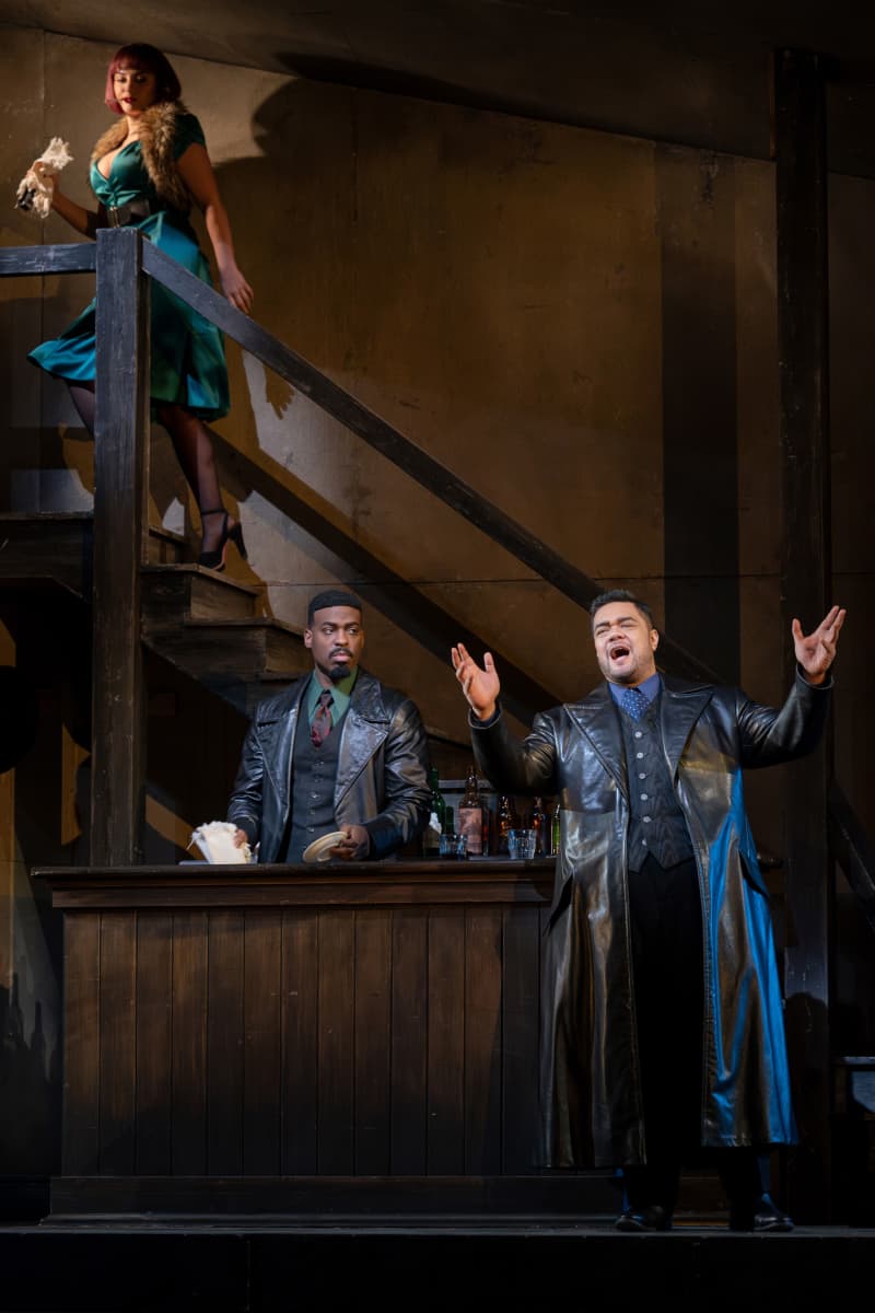 Pene stands on stage singing. He wears a large leather coat over a suit. Other actors glare at his character as he sings grandiosely.